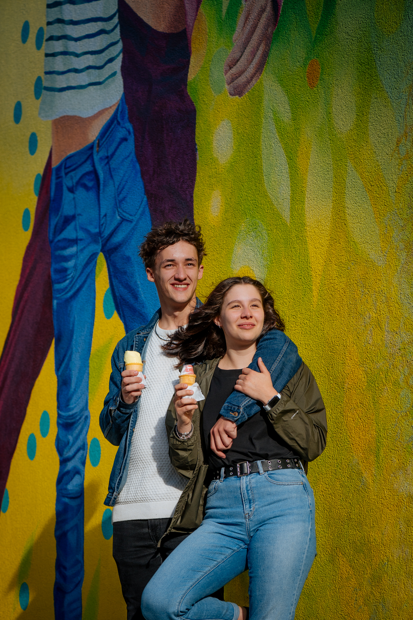 Un couple de jeunes adultes tiennent chacun un cornet de crème-glacée dans leur main. Le jeune garçon passe son bras sur les épaules de sa compagne. Tout deux sourient en regardant l'horizon. 